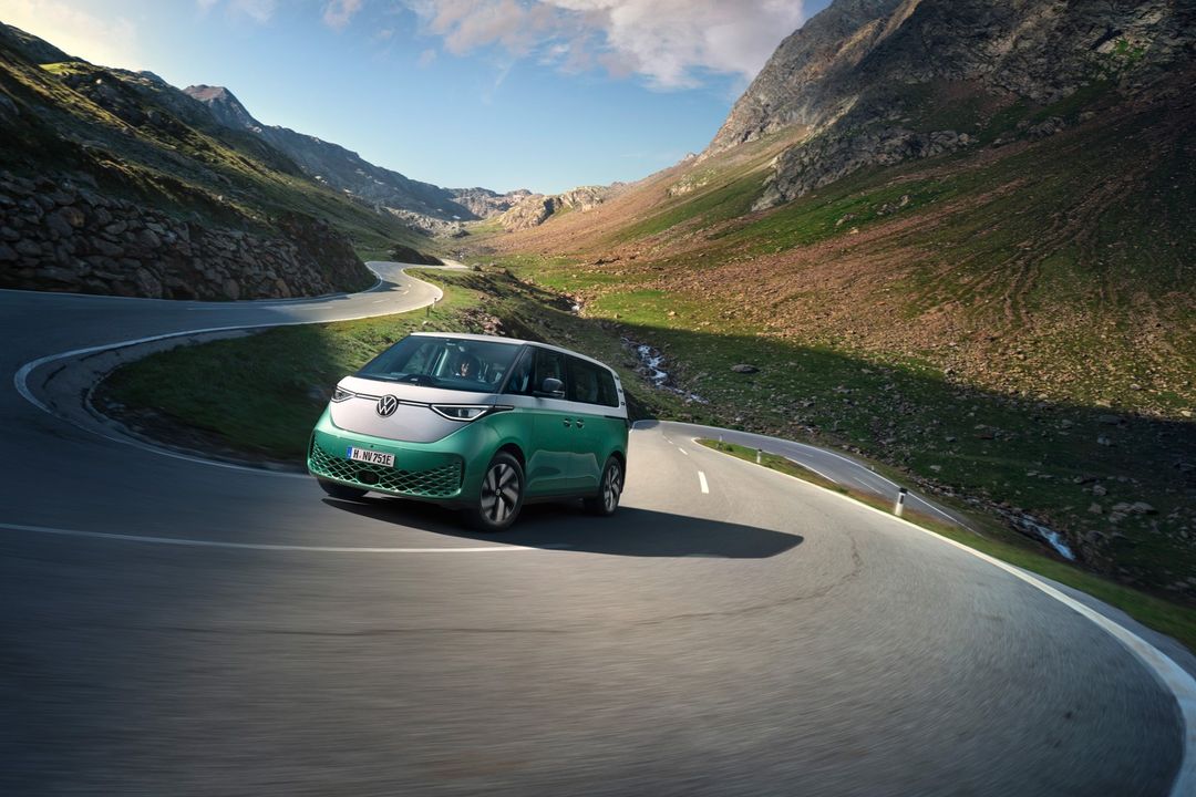 Mint green and white Volkswagen ID Buzz driving on winding road in mountains, blue sky, sunny day. Automotive advertisement.