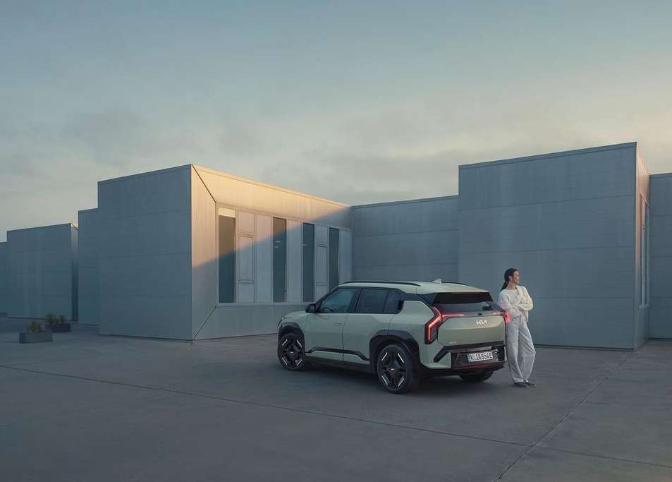 Modern electric SUV parked outside minimalist building with an adult female leaning against the car. Car is light green.
