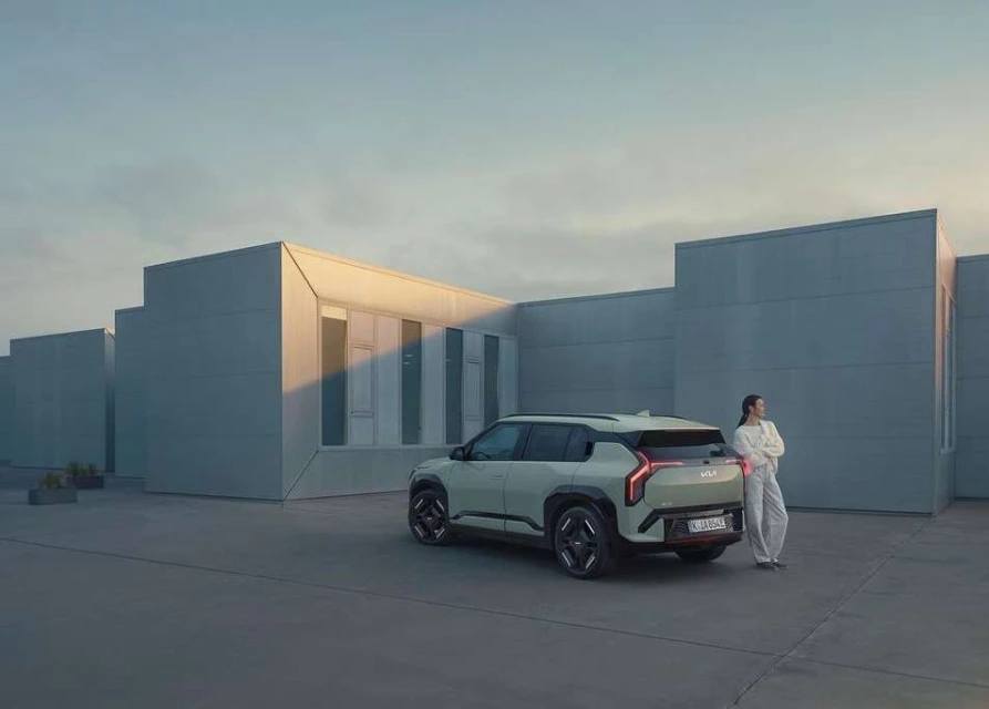 Modern electric SUV parked outside minimalist building with an adult female leaning against the car. Car is light green.