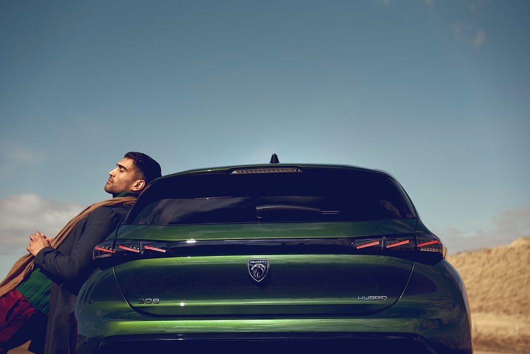 Profile of a man leaning against a green Peugeot 308 Hybrid car under a blue sky with tan grass visible in the background.