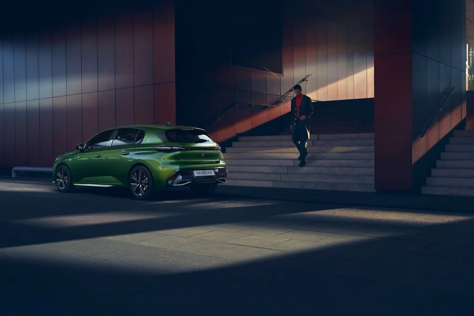 A green Peugeot car parked outside a modern building with a man walking down stairs on the right side of the frame.