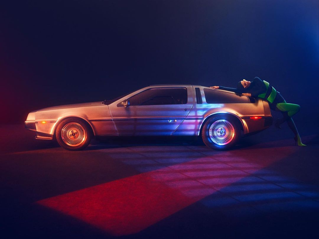 Profile view of a silver DeLorean automobile in a dark indoor setting with a woman in a green and black bodysuit leaning on the car.