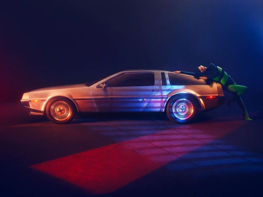 Profile view of a silver DeLorean automobile in a dark indoor setting with a woman in a green and black bodysuit leaning on the car.