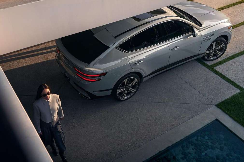 Woman in sunglasses and grey jacket standing near a light grey Genesis GV80 SUV parked on a concrete surface, captured from overhead.