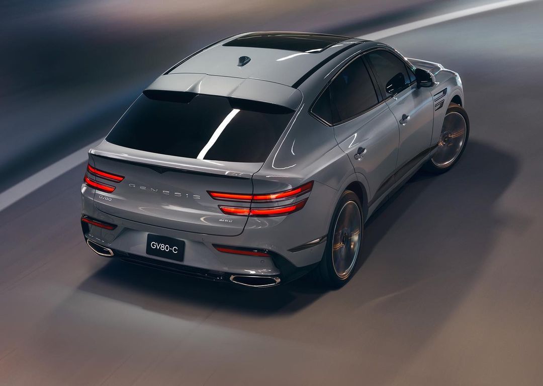 A gray Genesis GV80 Coupe drives on a blurred gray road, showcasing its sleek design and taillights in a nighttime setting.