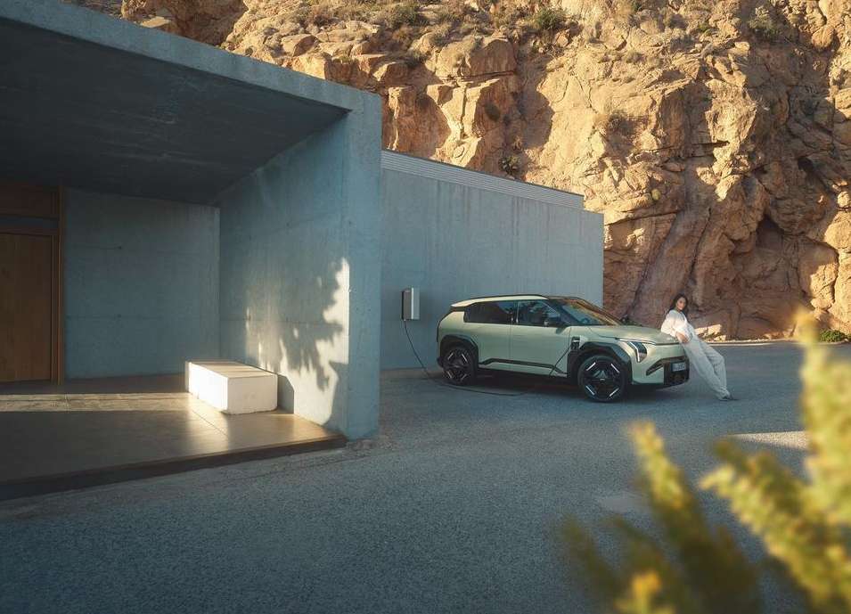 Electric SUV parked outside a modern concrete house with a woman leaning on the hood, against a desert mountain backdrop.
