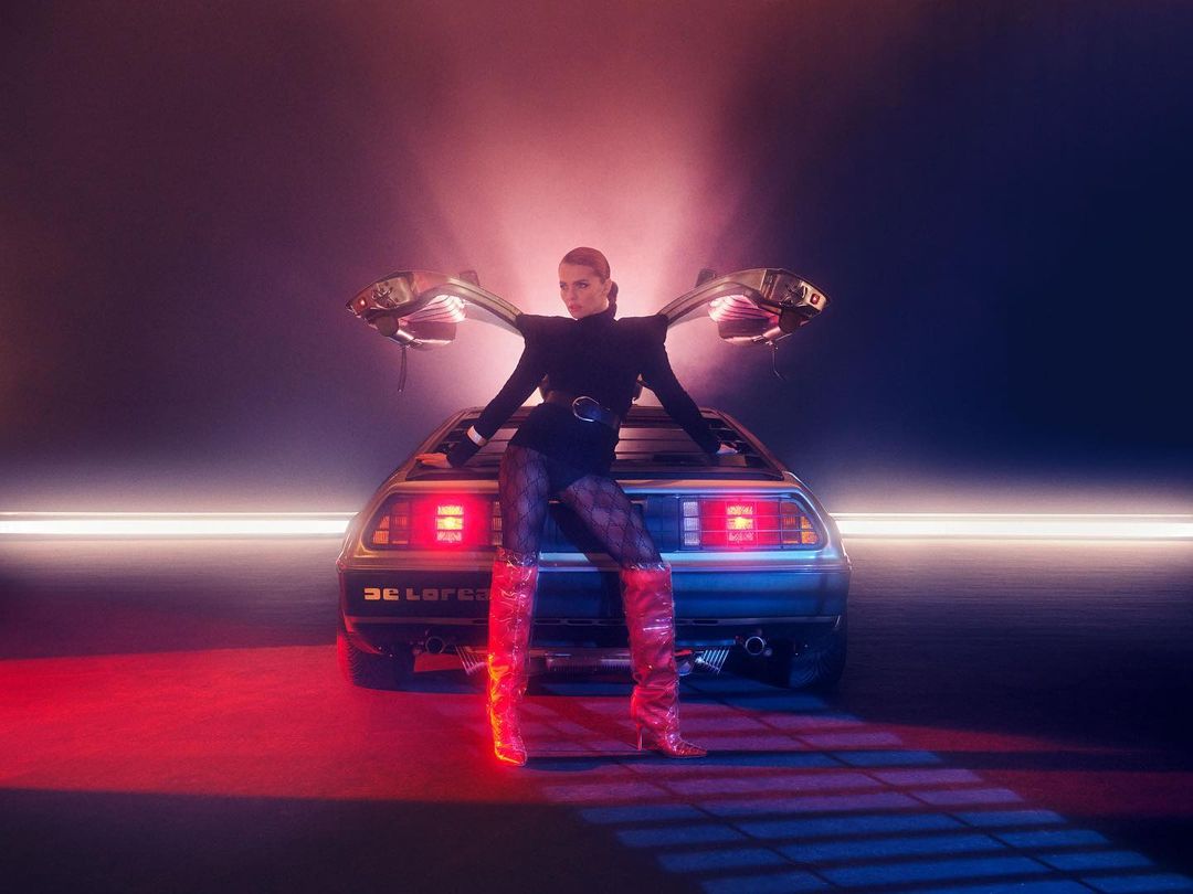 Woman in red boots poses with open-door DeLorean car in a studio with pink and blue lighting, creating a retro and futuristic vibe.