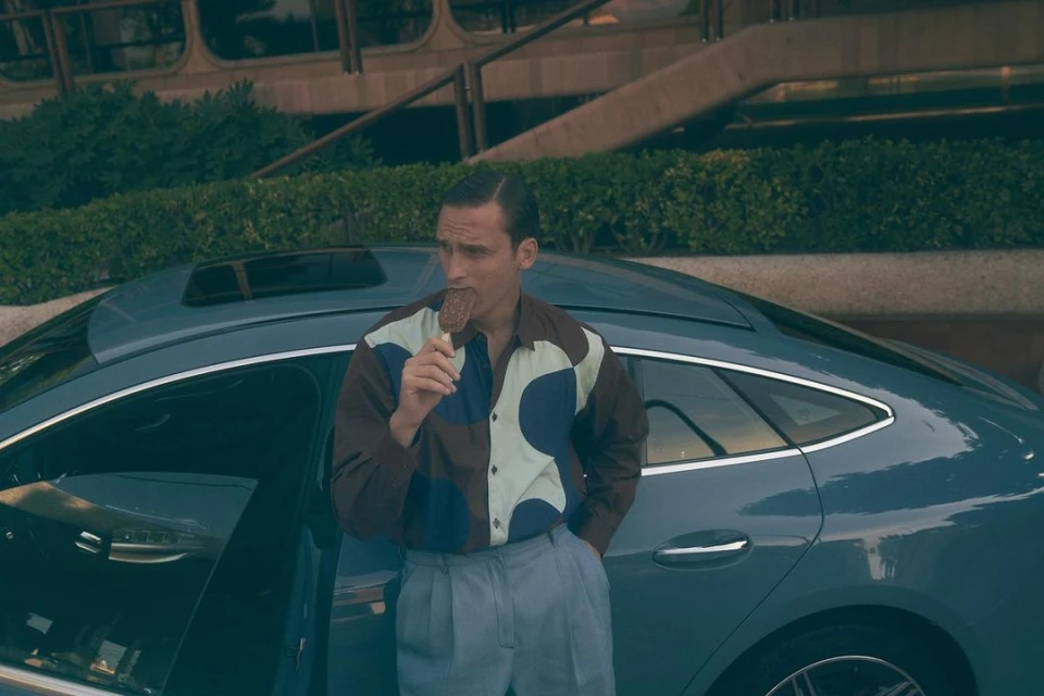A man in a patterned shirt and light trousers stands next to a blue car, enjoying an ice cream on a stick in an outdoor setting.