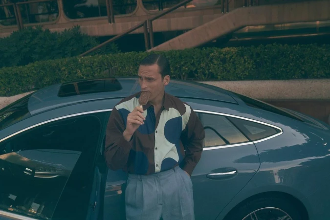A man in a patterned shirt and light trousers stands next to a blue car, enjoying an ice cream on a stick in an outdoor setting.