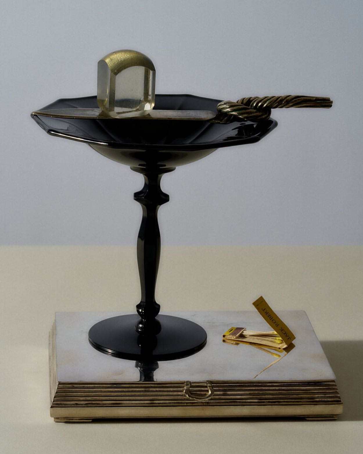 Still life photograph features a glass cube on a black pedestal, resting on a silver and gold decorative box, with a neutral backdrop.