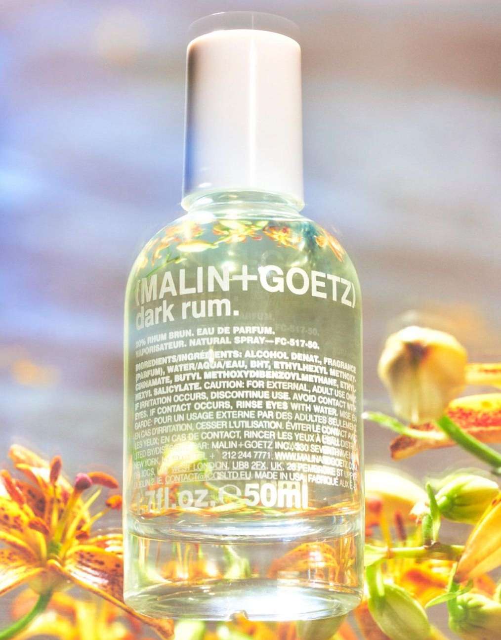Malin + Goetz 'Dark Rum' perfume bottle, clear glass with white cap, surrounded by yellow and orange flowers, beauty, fragrance.