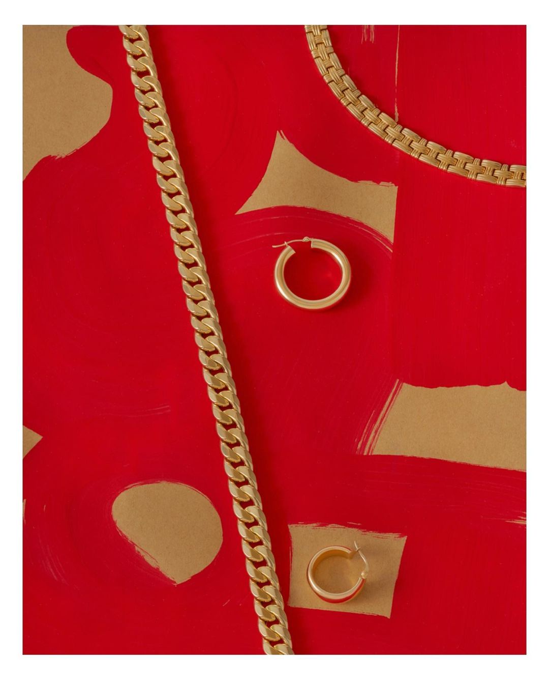 Still life flat lay featuring a gold cuban link chain, a gold box chain, and two gold hoop earrings displayed on a red surface with gold accents.