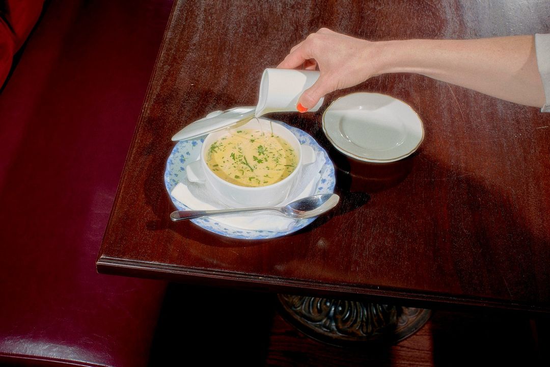 Overhead shot of a person pouring sauce over soup on a wooden table with a side plate, spoon, and cushioned seating.