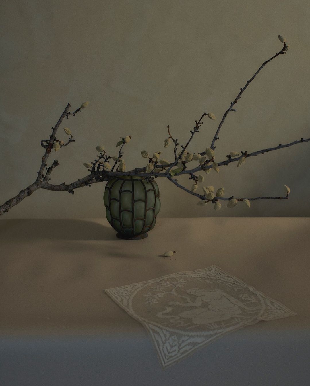 Still life photograph of a green vase with budding branches on a table, accompanied by a white embroidered cloth. Indoor setting.