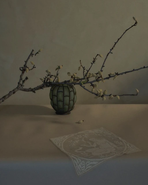 Still life photograph of a green vase with budding branches on a table, accompanied by a white embroidered cloth. Indoor setting.
