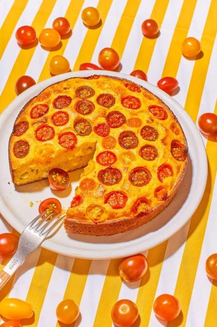 Overhead shot of a sliced tomato tart on a white plate, with a fork, resting on a yellow-and-white striped tablecloth, bright lighting.