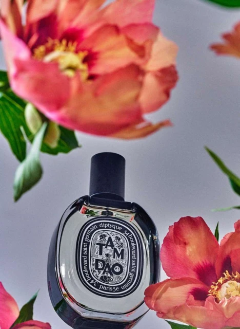 Elegant product shot of Diptyque's Tam Dao perfume bottle surrounded by pink and coral peony flowers with green leaves on white.