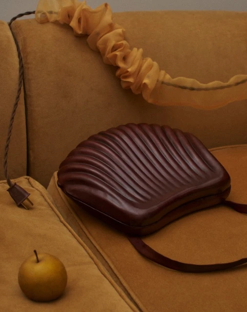 Close-up of a brown shell-shaped purse on a mustard yellow couch, with an apple, lamp cord, and decorative fabric.