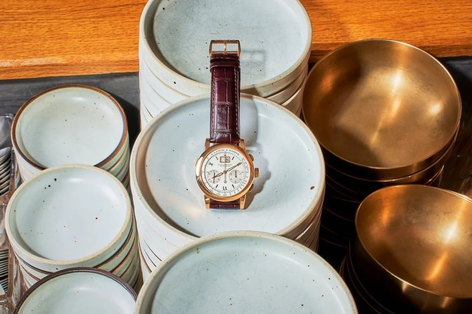 Luxury watch with gold finish and burgundy leather strap displayed on a stack of white ceramic plates with brass bowls in the background.