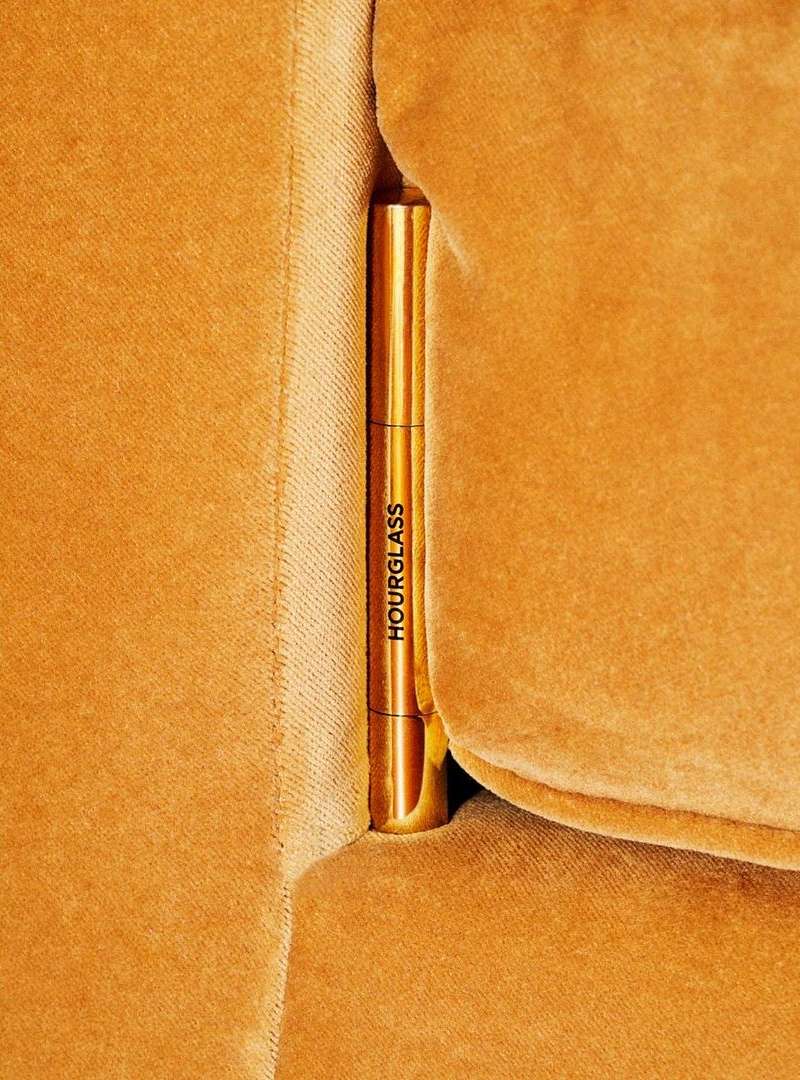Hourglass brand makeup product between the cushions of an orange velvet couch, in a close-up shot, with warm and golden lighting.
