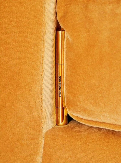 Hourglass brand makeup product between the cushions of an orange velvet couch, in a close-up shot, with warm and golden lighting.
