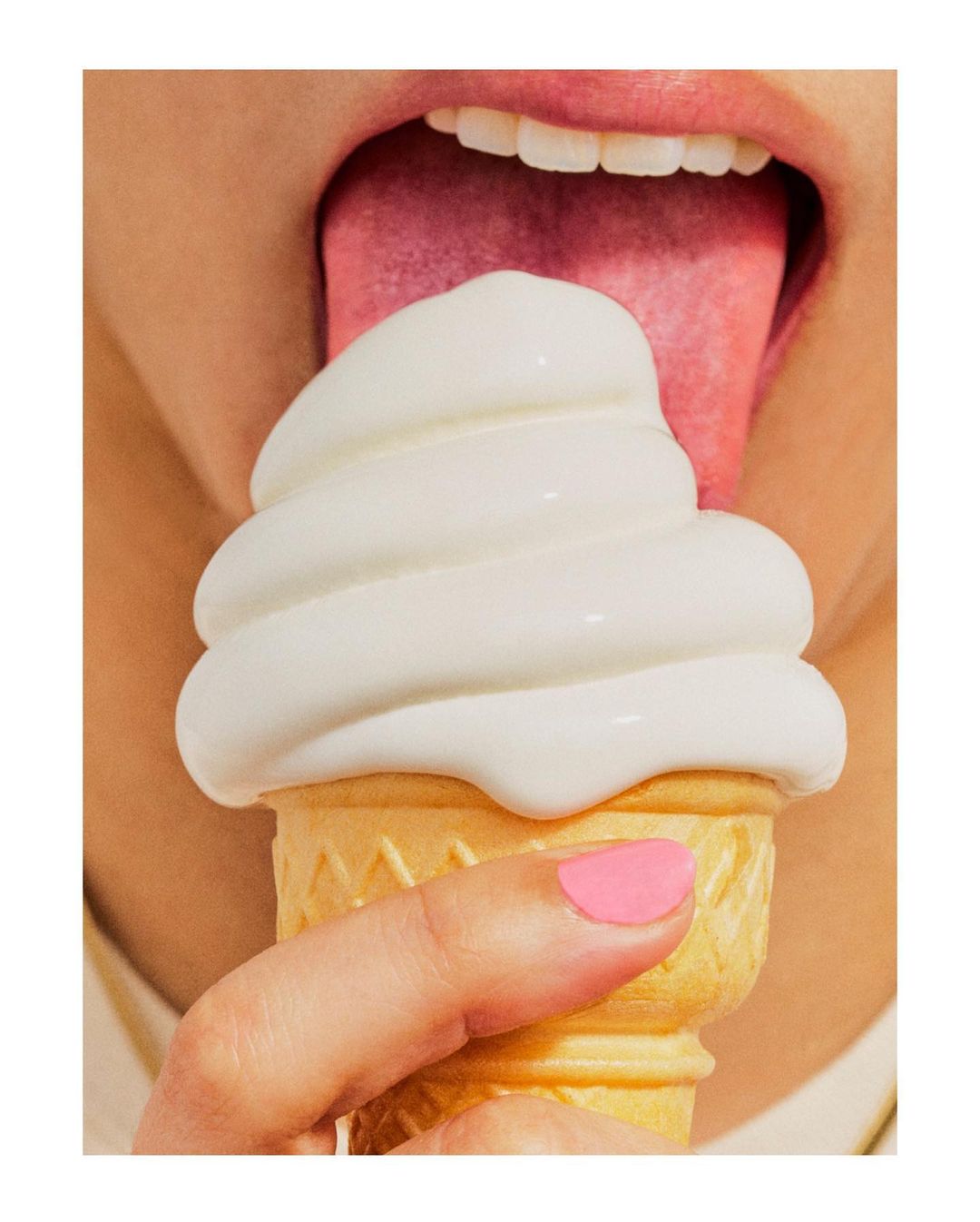 Close-up shot of a woman with an ice cream cone being licked by her tongue. The ice cream is white, and the woman has pink nail polish.