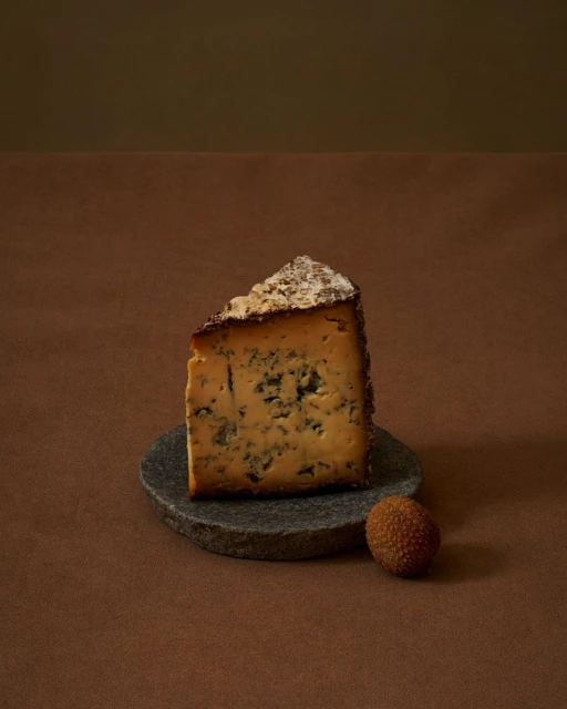 Studio shot of a wedge of blue cheese on a stone coaster, accompanied by a lychee fruit, all set against a gradient brown backdrop.
