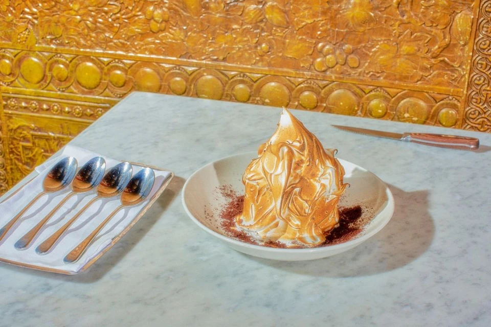 A dessert with toasted meringue on a white plate, with spoons, a knife, and gold decorative background on a marble table.