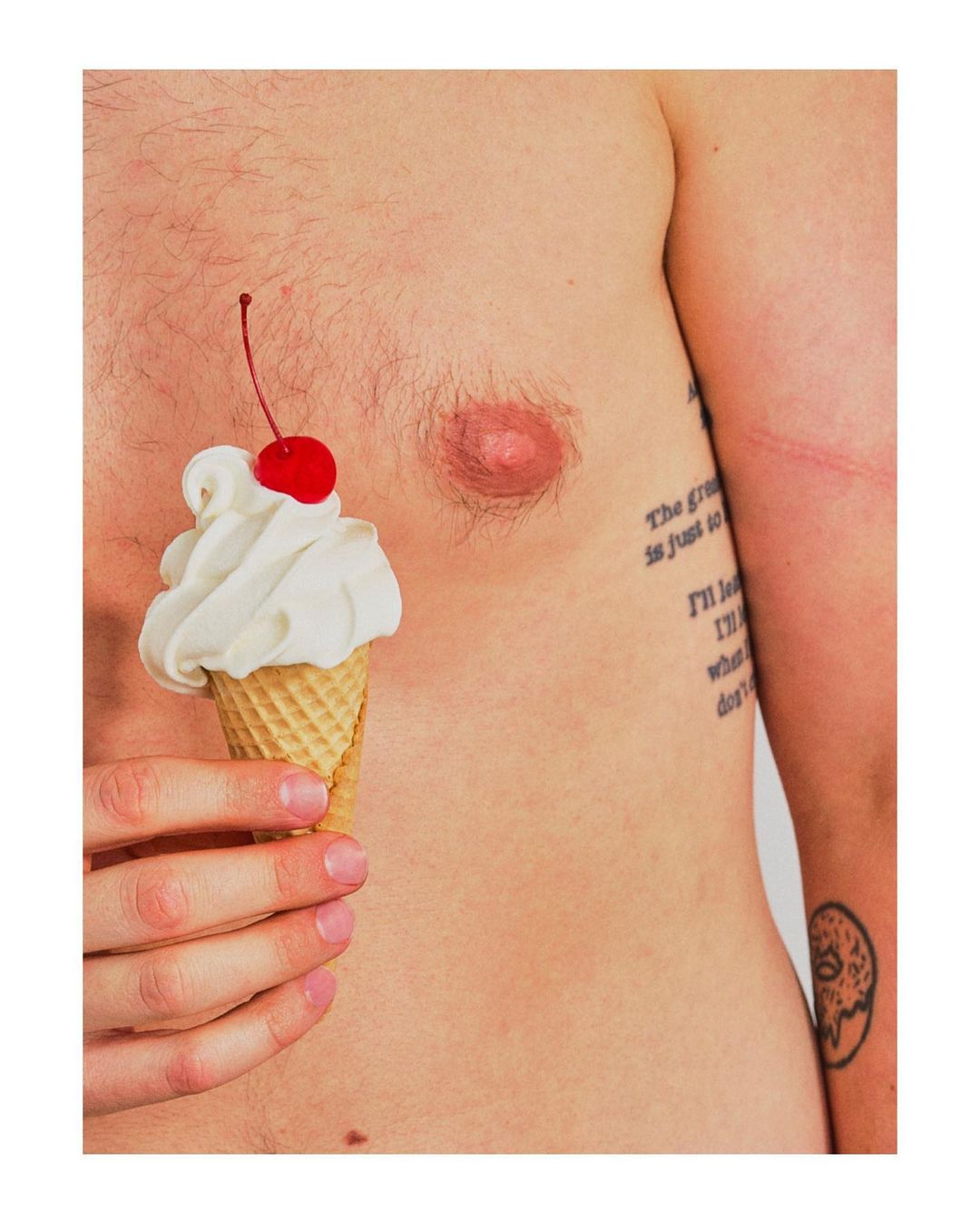 Man holding ice cream cone with cherry on his bare chest in a close-up, studio shot with bright, even lighting and white background.