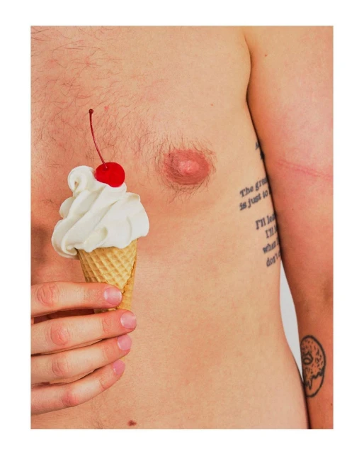 Man holding ice cream cone with cherry on his bare chest in a close-up, studio shot with bright, even lighting and white background.