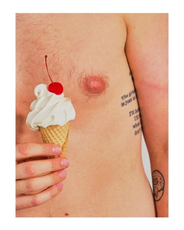 Man holding ice cream cone with cherry on his bare chest in a close-up, studio shot with bright, even lighting and white background.