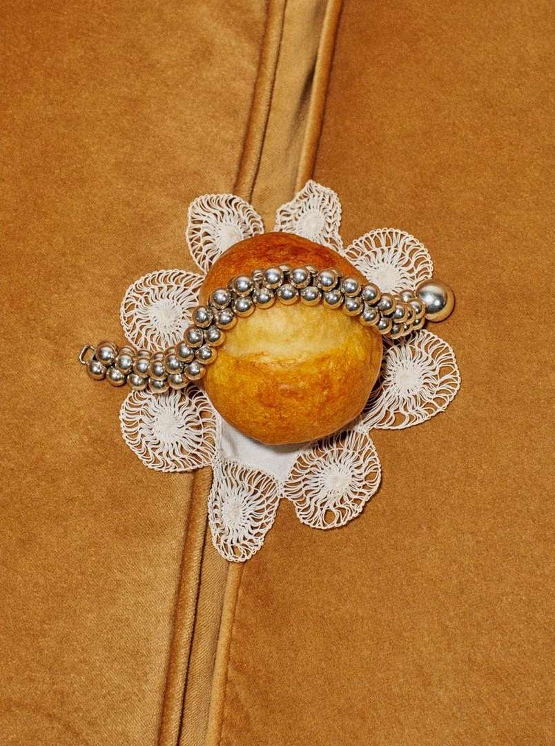Overhead shot of an apple on a white lace doily, adorned with a silver beaded bracelet, set against a brown fabric background.