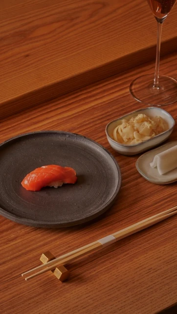 Overhead shot of salmon nigiri on a black plate with chopsticks and condiments on a wooden table near a glass of rose wine.