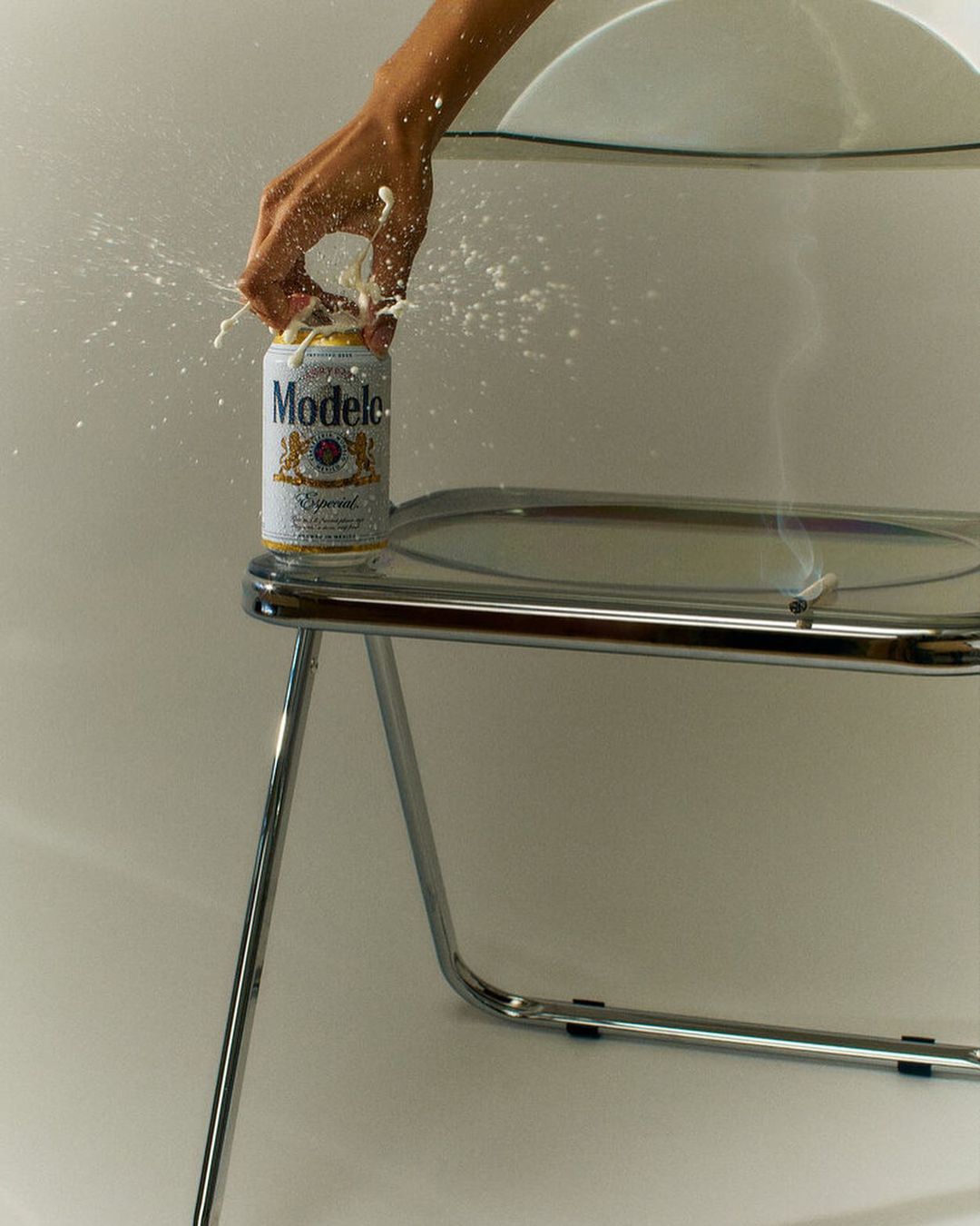 A can of Modelo beer is being opened, with liquid splashing as a tan-skinned hand holds it, against a white backdrop with a silver chair.