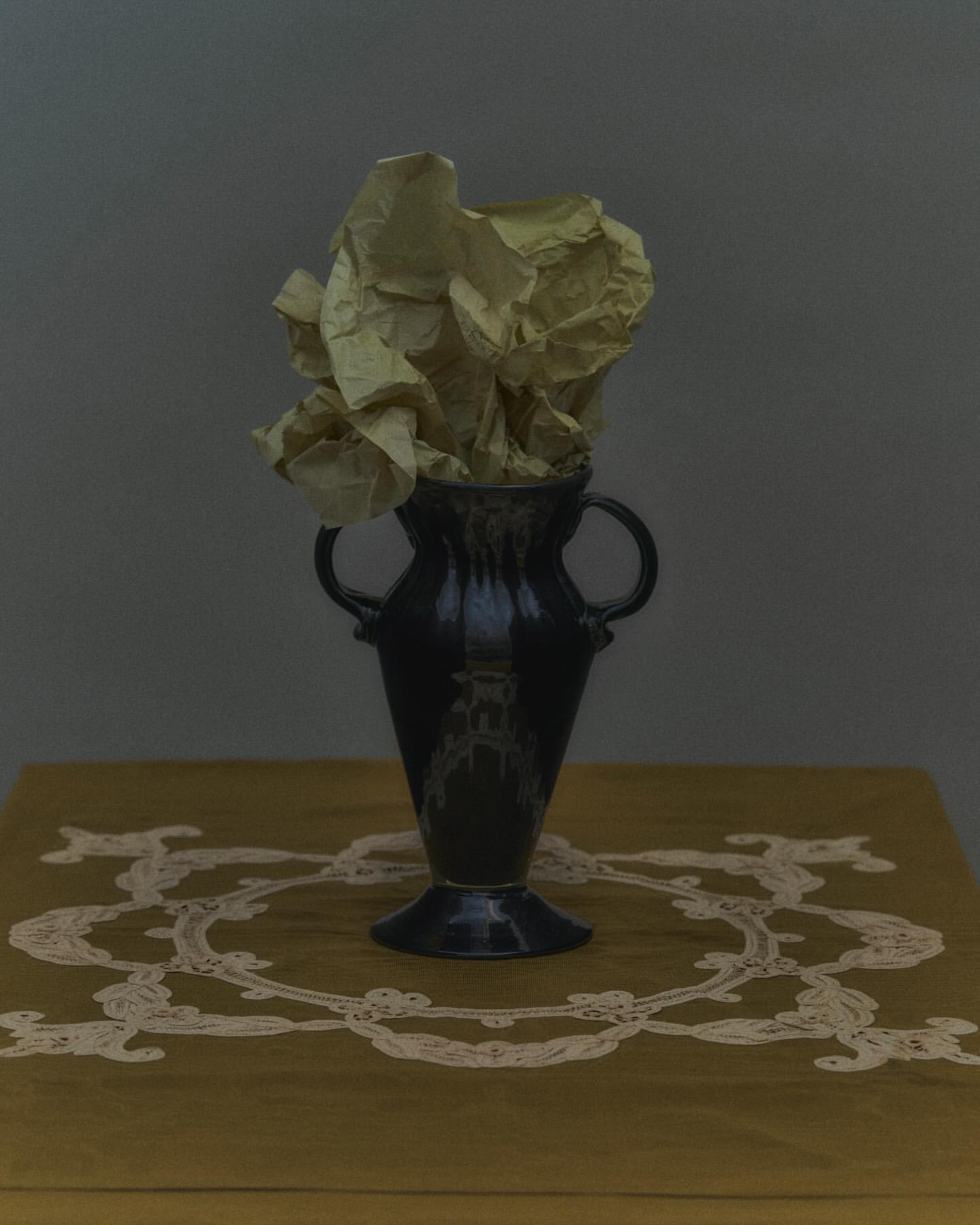 Still life photograph featuring a dark-glazed vase filled with crumpled yellow paper on a yellow table with white lace trim against gray background.