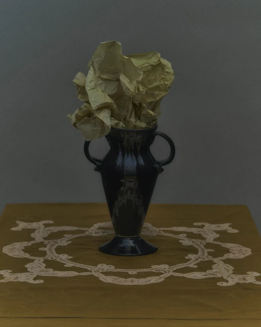 Still life photograph featuring a dark-glazed vase filled with crumpled yellow paper on a yellow table with white lace trim against gray background.