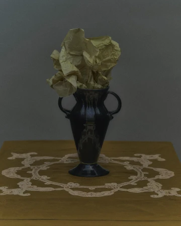 Still life photograph featuring a dark-glazed vase filled with crumpled yellow paper on a yellow table with white lace trim against gray background.