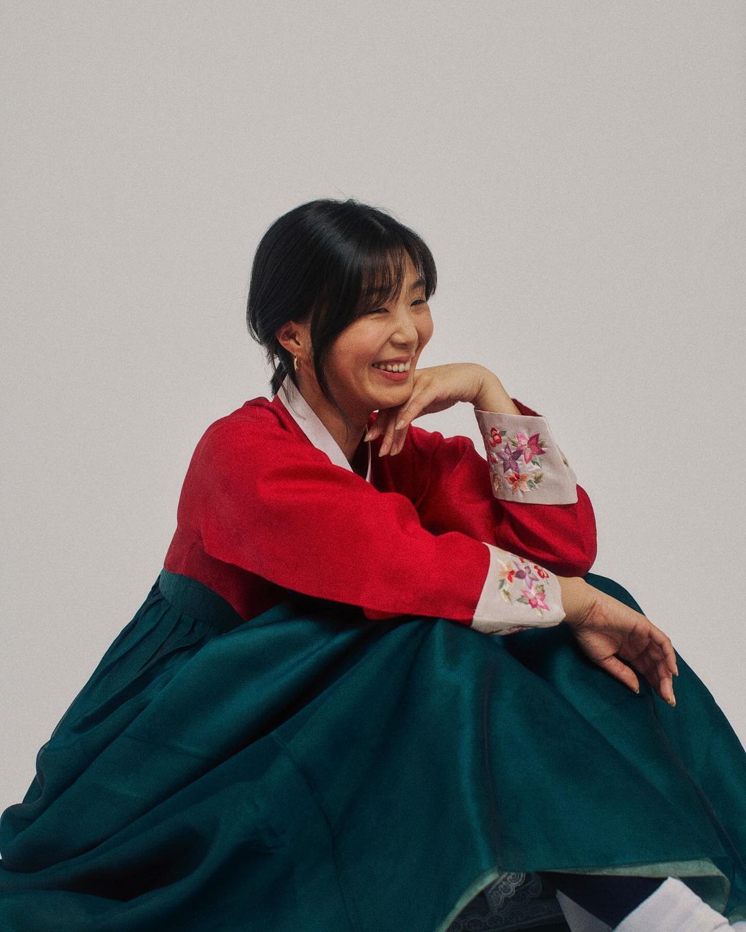 Smiling Asian woman in traditional Korean hanbok dress with red top and green skirt, posing in a bright photo studio.