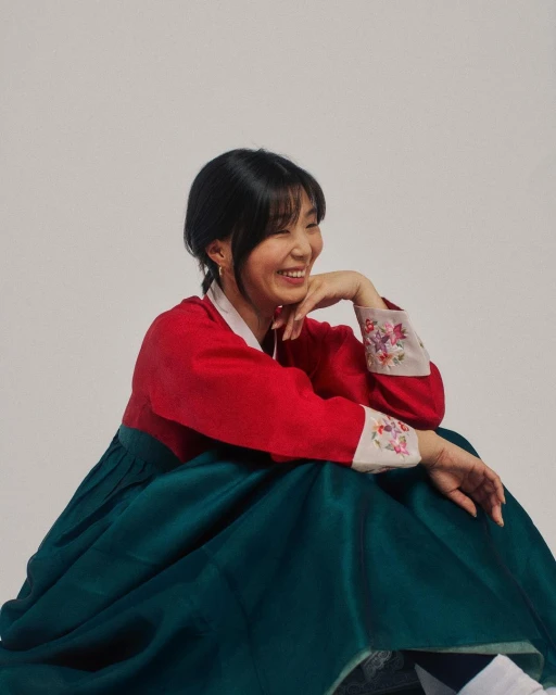 Smiling Asian woman in traditional Korean hanbok dress with red top and green skirt, posing in a bright photo studio.