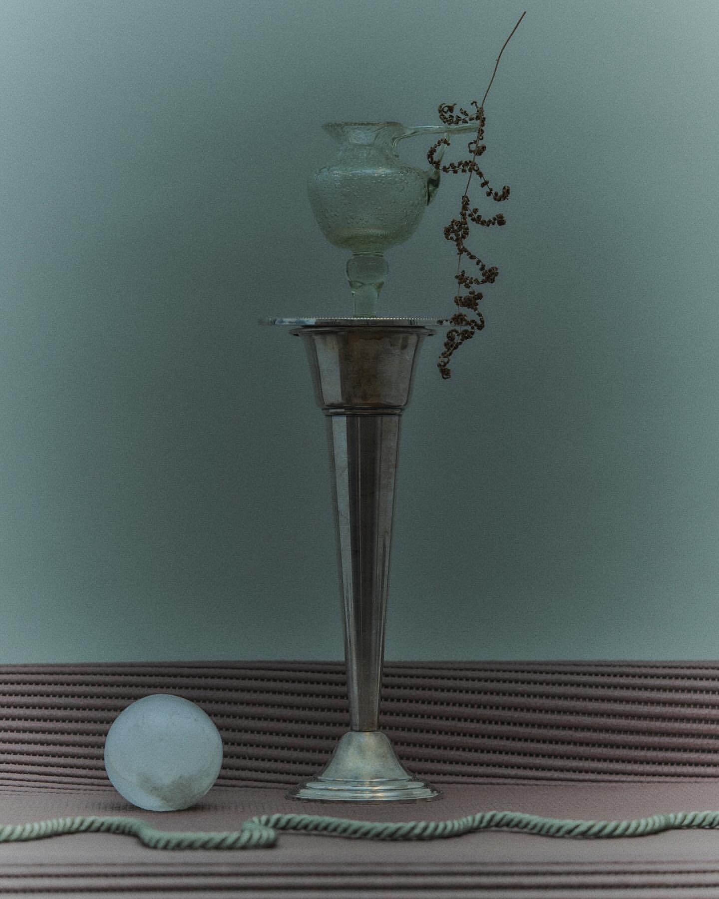 Silver vase with a clear glass vase on top and decorative strands, next to a rope and a white sphere, creates a unique still life.