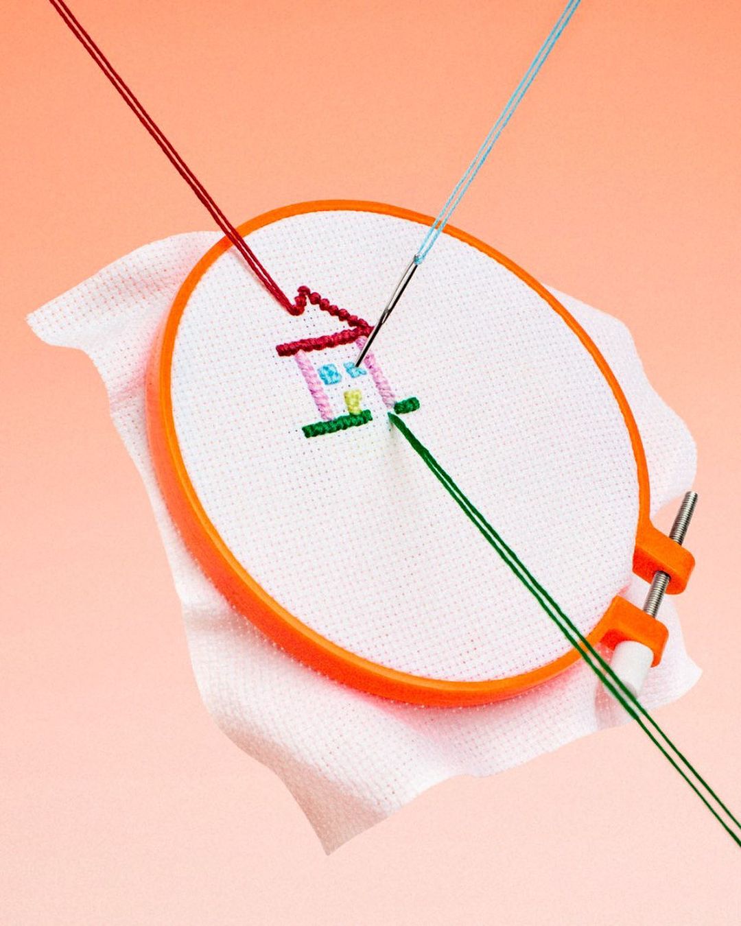Cross-stitch embroidery project in progress, showcasing colorful threads forming a house design on white fabric within an orange hoop.