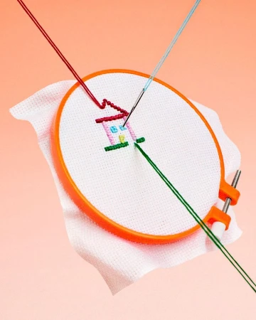 Cross-stitch embroidery project in progress, showcasing colorful threads forming a house design on white fabric within an orange hoop.