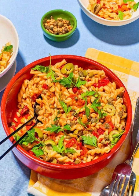 Overhead shot of a red bowl of fusilli pasta mixed with olives, capers, tomatoes and parsley on a blue tablecloth.