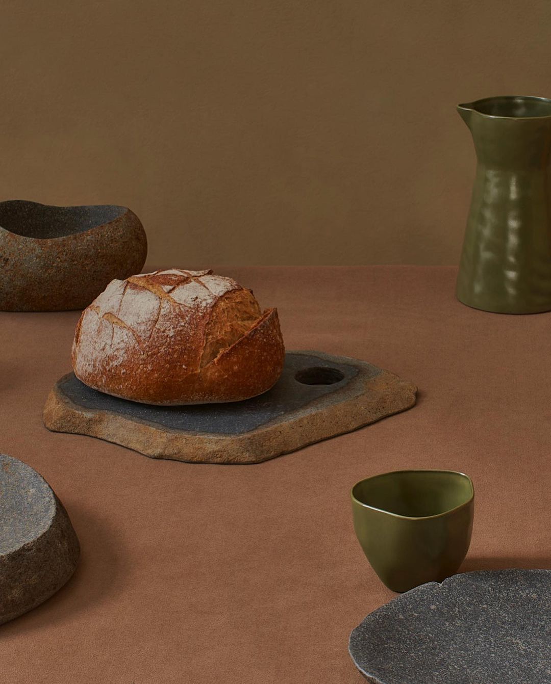 Artisanal bread sits on a stone cutting board amid a collection of stone bowls and a green ceramic pitcher against a brown backdrop.