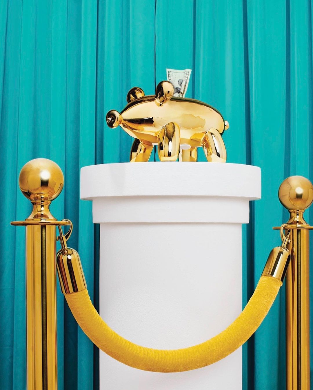 Close-up of a golden piggy bank on a white column, guarded by gold stanchions with yellow rope, set against a vibrant turquoise curtain backdrop.