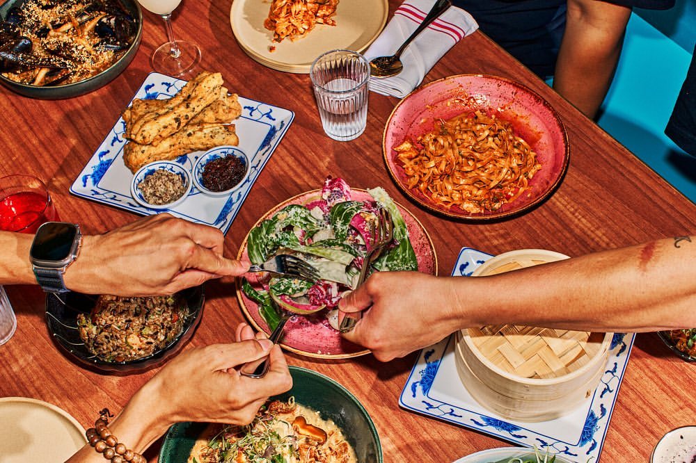Overhead shot of diverse food dishes on a wooden table surrounded by hands using cutlery in a restaurant setting with mixed lighting.