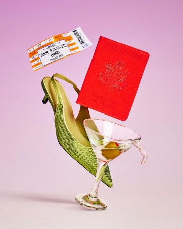 Still life of an open US passport, green high-heeled shoe, martini glass, and concert ticket floating in front of a pink background.