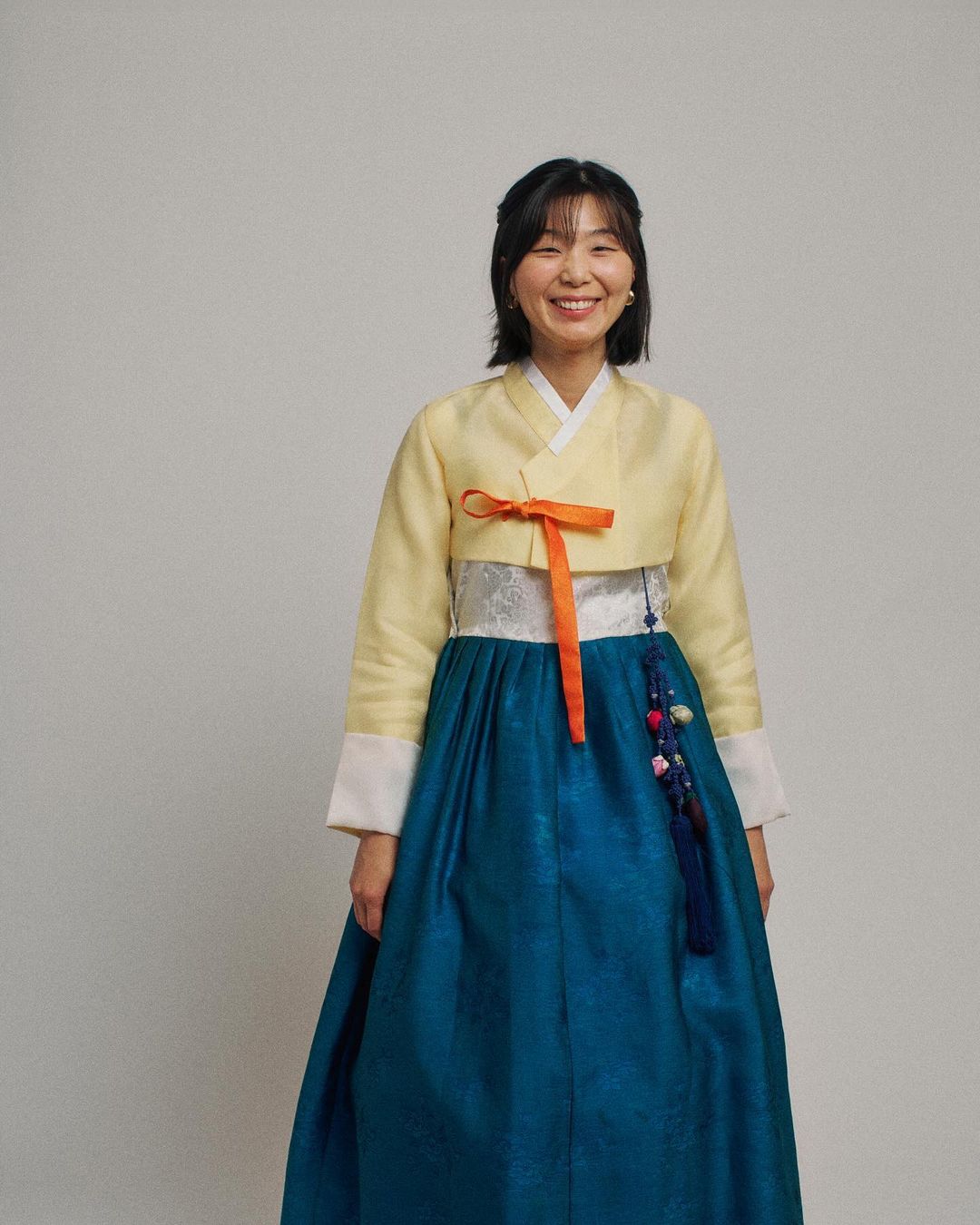 Smiling Asian woman in traditional yellow and blue Hanbok dress, posing in a studio with a light background. Portrait of a woman.