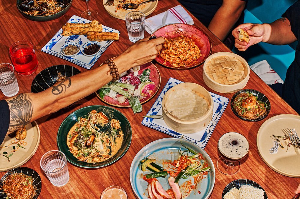 Overhead shot of a wooden table covered with colorful plates of Asian cuisine; hands reach into frame, with drinks, bowls, and bamboo steamers.