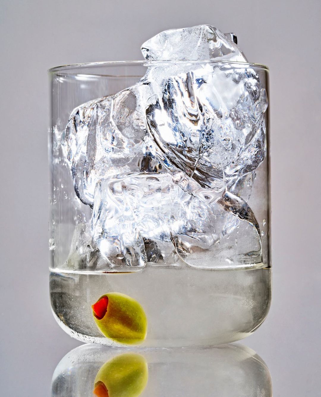 Close-up of a clear glass tumbler filled with large ice cubes and a single green olive resting at the bottom